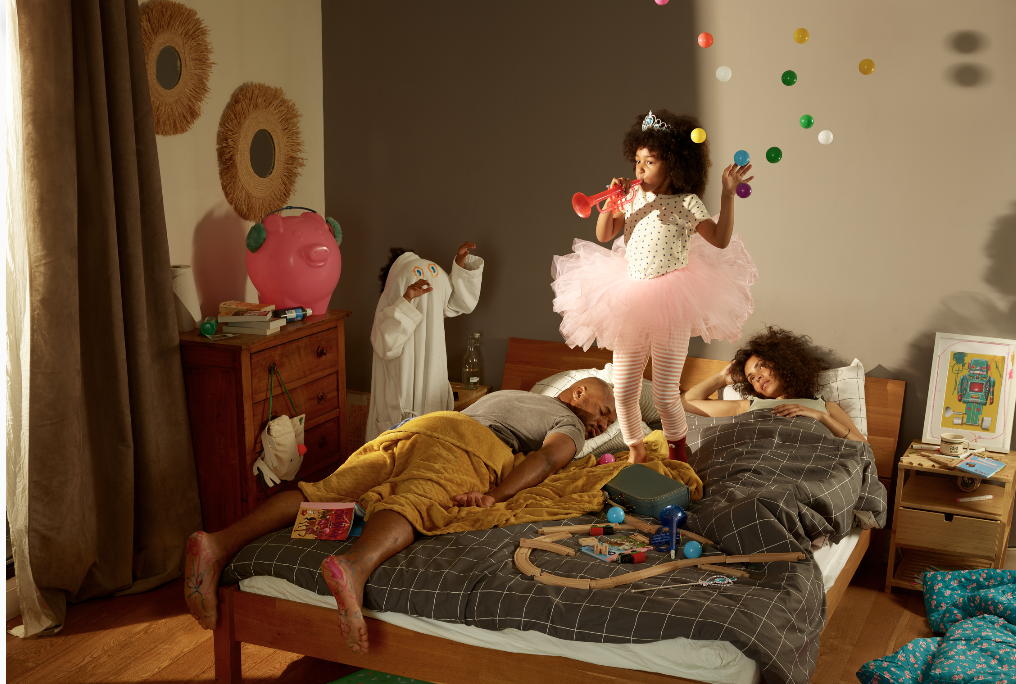 Couple allongé sur un lit, épuisés, avec leur enfant sautant sur le lit.